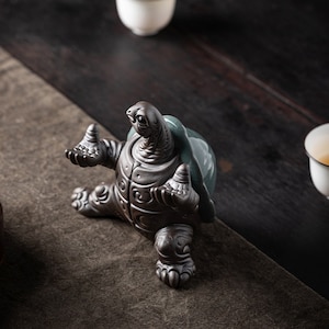 May include: A ceramic turtle figurine with a green shell and brown body. The turtle is sitting on its hind legs with its front legs outstretched. The figurine is a decorative piece for a home or office.