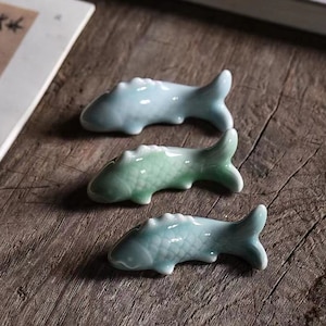 May include: Three ceramic fish-shaped chopsticks rest on a wooden surface. The chopsticks are glazed in shades of blue and green.