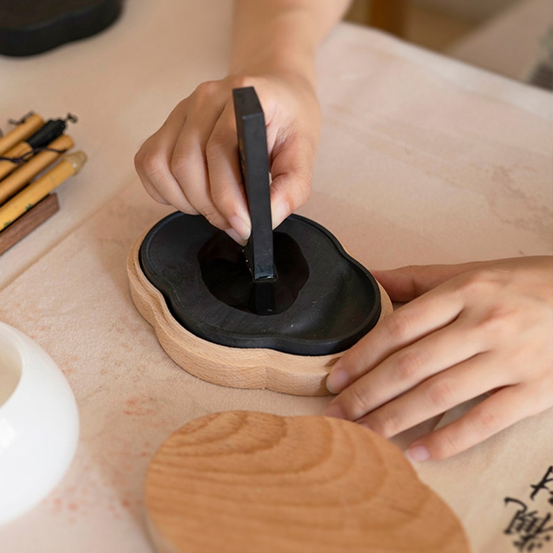 May include: A person grinding an ink stick in a black ink stone, which is set in a wooden holder. The ink stone is shaped like a cloud and has a dark brown wooden base.