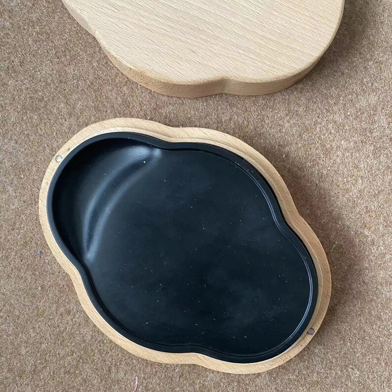 May include: A wooden box with a black, smooth, oval-shaped insert. The box is shaped like a cloud and has a light wood finish.