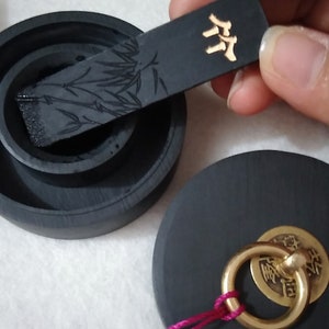 May include: A black ink stone with a lid and a small black ink stick with a bamboo design. The ink stick has a gold label with the word "竹" written on it. The ink stone has a small brass ring with a gold disc that has Chinese characters on it.
