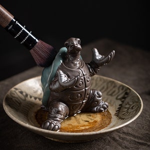 May include: A ceramic turtle tea pet with a detailed design, holding a tea brush. The turtle is sitting in a brown and white ceramic dish.