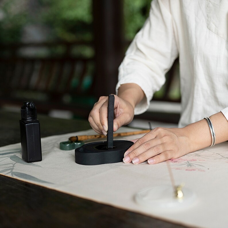 May include: A person using a black ink stick on a black ink stone. The ink stone is on a white surface with a green and brown brush next to it.