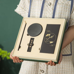 May include: A black gift box with a green border contains a black ink stone, a black ink stick, a black ink slab, and a black calligraphy brush. The ink stick has gold lettering that reads "品行端" in Chinese.