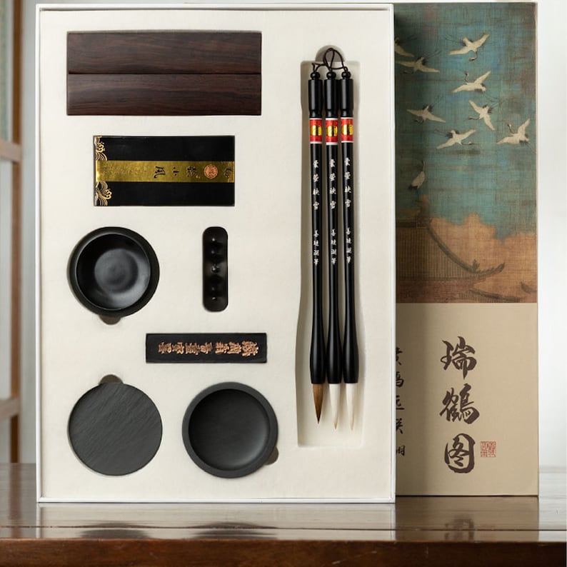 May include: A white box with a black ink stone, three black calligraphy brushes, a black ink stick, and two black inkwells. The box also includes a wooden block with the text "書道" in black. The box is open and the contents are visible. The box is sitting on a wooden table.