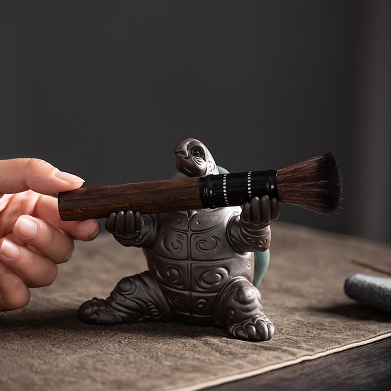 May include: A ceramic turtle figurine holding a brown and black brush. The turtle has a detailed shell and is sitting on a brown surface.