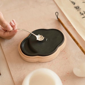 May include: A black ink stone with a wooden base, a silver spoon, and a wooden calligraphy brush. The ink stone is shaped like a cloud and has a smooth surface. The spoon is used to stir the ink and the brush is used to write calligraphy.