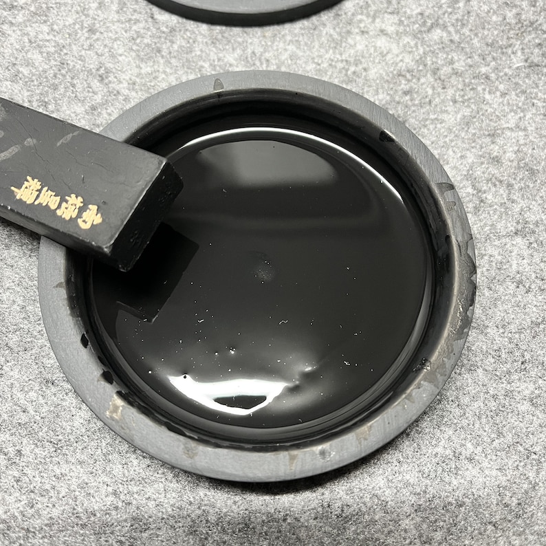 May include: A black ink stone with a black ink stick resting on the edge. The ink stone is filled with black ink.