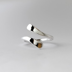 Könnte beinhalten: Ein silberfarbener Ring mit einem einzigartigen, fließenden Design. Der Ring umschließt den Finger mit zwei gebogenen, polierten Bändern, die sich überlappen. Die glatte, reflektierende Oberfläche des Rings fängt das Licht ein und erzeugt Glanzlichter und Schatten.