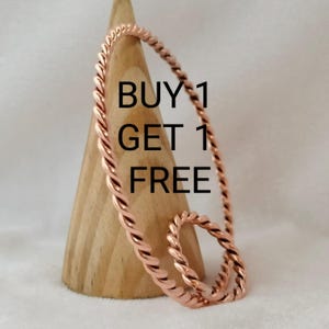 Op de afbeelding: Twee koperen armbanden met een gedraaid touwontwerp. De armbanden worden gepresenteerd op een houten kegel. De tekst "BUY 1 GET 1 FREE" is zichtbaar in de afbeelding.