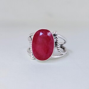 Könnte beinhalten: Ein silberner Ring mit einem großen, ovalen, rubinroten Edelstein. Der Ring hat ein Doppelband-Design mit seilartigen Details an den Seiten. Der Edelstein ist der Mittelpunkt und zeigt seine leuchtende Farbe.