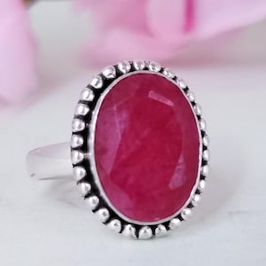 May include: A silver ring with a large, oval-cut, deep red gemstone. The gemstone is framed by a beaded border. The ring is set against a soft, blurred background, highlighting the jewelry's design and colour.