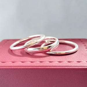 May include: Three silver rings with a polished finish are arranged on a red, textured surface. The rings have a simple, elegant design. The rings are stacked together, creating a layered effect. The background is a soft grey.