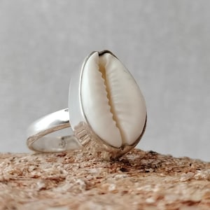 Puede incluir: Un anillo de plata con una concha de cauri blanca engastada en el centro. El anillo está sobre una superficie de madera.