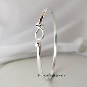 Puede incluir: Un brazalete plateado con un diseño abierto. El brazalete presenta un detalle en forma de nudo y un extremo cuadrado. La joya se exhibe sobre una superficie blanca. El texto "Dishgemsandjewelry" es visible.