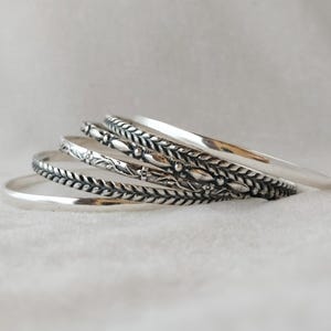 Puede incluir: Cinco pulseras de plata con diferentes diseños grabados. Las pulseras están apiladas unas sobre otras.