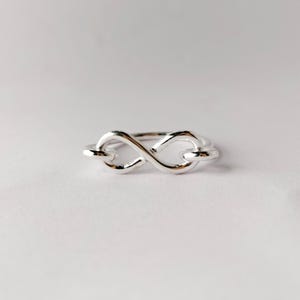 May include: A silver ring featuring an infinity symbol design. The ring is made of a polished metal and is displayed on a white background. The infinity symbol is a continuous loop, representing eternity and endless possibilities.