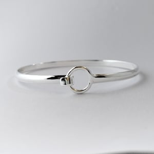 May include: A silver bangle bracelet with a circular clasp. The bracelet is made of polished metal and has a simple, elegant design. The clasp is a small, rectangular piece that connects to a circular loop. The bracelet is shown on a white background.