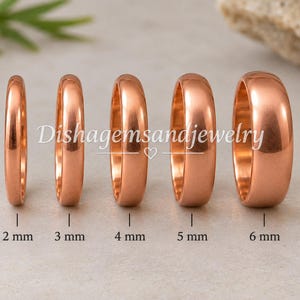 Op de afbeelding: Vijf gepolijste koperkleurige ringen op een rij, oplopend in breedte van 2 mm tot 6 mm. De tekst Dishagemsandjewelry staat gecentreerd over de ringen. Elke ring is eronder gelabeld met de specifieke breedtemaat in millimeters.