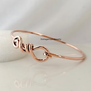 May include: A rose gold bangle bracelet with a unique knot design. The bracelet features a looped knot detail on one side and a smooth, slender band. The text "Dishgemsandjewelry" is visible on the right side.