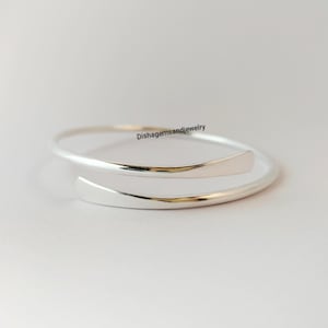 Könnte beinhalten: Ein silberfarbenes Armreif-Armband mit offenem Design. Das Armband hat eine glatte, polierte Oberfläche und hat an jeder Seite ein gebogenes, flaches Ende. Der Text "Dishgemsandjewelry" ist auf dem Bild sichtbar.