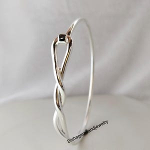 Puede incluir: Un brazalete plateado con un diseño retorcido. La pulsera presenta un lazo y una sección trenzada, creando un aspecto elegante y moderno. La joya se exhibe sobre un fondo blanco.