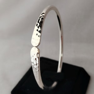 May include: A silver-colored open cuff bracelet with a hammered texture on the ends. The bracelet is displayed on a black surface against a neutral background. The ends of the bracelet are slightly flattened and textured.