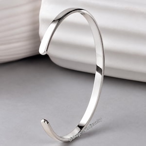 May include: A polished silver-tone open cuff bracelet with a smooth, rounded surface and tapered ends. The metal has a reflective finish. The text Dishagemsandjewelry is printed in small black letters near the bottom of the bracelet.