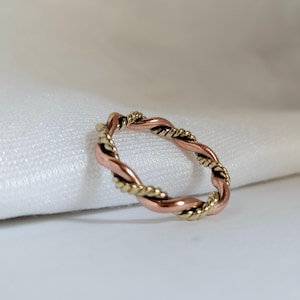 May include: A twisted ring featuring a combination of copper and gold-colored metal. The ring has a braided design, with the two metal colors intertwined to create a textured appearance. The ring is set against a white background.