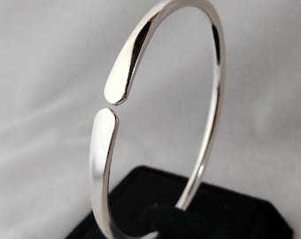 Könnte beinhalten: Ein silbernes Armreif-Armband mit einem einfachen, offenen Design. Das Armband besteht aus einem dicken, runden Draht und hat eine glatte, polierte Oberfläche.