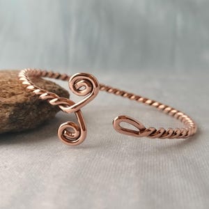 May include: A copper-colored metal cuff bracelet featuring a twisted wire design. The bracelet has an open-ended style with two prominent spiral-shaped terminals at one end and a single loop-shaped terminal at the other end.