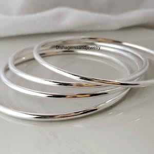 Może przedstawiać: Cztery srebrne bransoletki ułożone na jasnej powierzchni. Bransoletki są okrągłe i mają gładkie, odbijające światło wykończenie. Tekst "Dishgemsandjewelry" jest widoczny na zdjęciu.