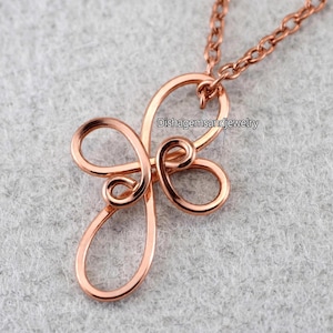 Peut inclure: Un collier avec un pendentif en forme de croix en fil métallique de couleur or rose, composé de boucles continues. La surface du métal est lisse et brillante. Le texte Dishagemsandjewelry est visible sur le pendentif.