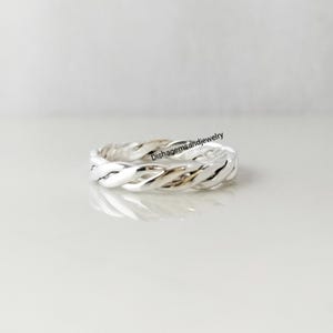 May include: A silver-colored ring with a braided design. The ring is made of a shiny metal and has a textured surface. The ring is displayed on a white surface. The text "Dishagemsandjewelry" is visible on the ring.