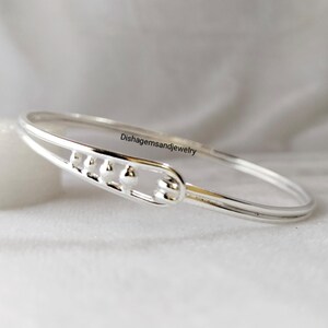 May include: A silver bangle bracelet with a decorative section featuring small, round embellishments. The bracelet has a smooth, polished finish and is displayed against a soft, white background. The text "Dishagemsandjewelry" is visible on the bracelet.