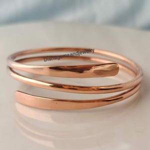 May include: A rose gold-colored metal bangle bracelet featuring a triple-wrap spiral design with smooth, flat, and polished surfaces. The text Dishagemsandjewelry is printed across the top of the bracelet in a dark, thin, sans-serif font.