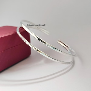 May include: Two silver-colored, hammered-texture bangles. The open-ended bracelets are displayed on a light surface next to a maroon box. The text "Dishgemsandjewelry" is visible.