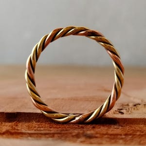 Könnte beinhalten: Ein gedrehter Ring aus drei verschiedenen Metallen: Gold, Kupfer und Silber. Der Ring ist schlicht gehalten und hat eine glatte Oberfläche.