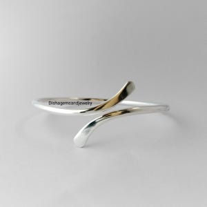 Könnte beinhalten: Ein silberfarbenes Armband mit minimalistischem Design. Das Armband hat ein gebogenes, offenes Design mit zwei sich überlappenden Enden. Die Oberfläche ist glatt und reflektierend, mit dem Text "Dishgemsandjewelry" sichtbar.