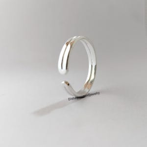 May include: A silver adjustable ring with a smooth, curved design. The ring has an open design, allowing for size adjustment. The ring is made of a shiny, reflective metal, set against a neutral background.
