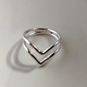 Peut inclure: Une bague argentée avec un design en forme de double V. La bague a une surface polie et réfléchissante, et les deux formes en V sont parallèles. La bague est probablement en argent sterling ou en métal similaire.
