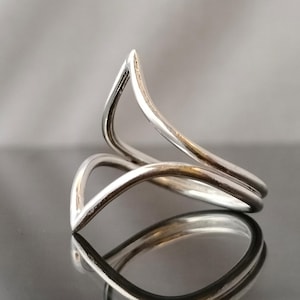 Könnte beinhalten: Ein silberner Ring mit einem einzigartigen, abstrakten Design. Der Ring besteht aus zwei gebogenen Bändern, die sich kreuzen und eine spitze Form bilden.