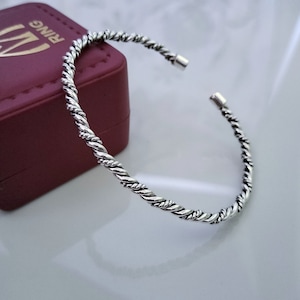 Puede incluir: Un brazalete de plata trenzado. El brazalete está abierto y tiene un diseño sencillo.