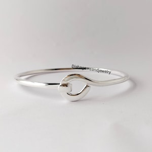 May include: A silver-colored bangle bracelet with a hook-and-eye closure. The bracelet is simple and elegant, with a smooth, polished finish. The clasp is a heart-shaped design. The text "Dishagemsandjewelry" is visible on the image.