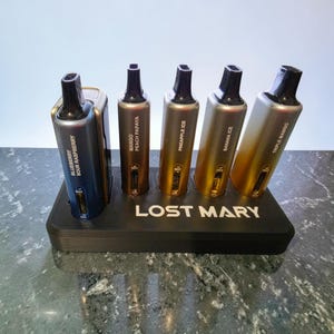 May include: Five Lost Mary disposable vape devices arranged in a black display stand. The devices feature gradient metallic finishes in blue, bronze, and gold, labeled with flavors including Blueberry Sour Raspberry, Mango Peach Papaya, Pineapple Ice, Banana Ice, and Triple Mango.
