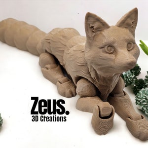 May include: A tan-coloured, articulated fox figurine. The fox is lying down with its legs extended and tail curved. The text "Zeus. 3D Creations" is printed in black on the lower left. The fox is made of a material that appears to have a wood grain texture.