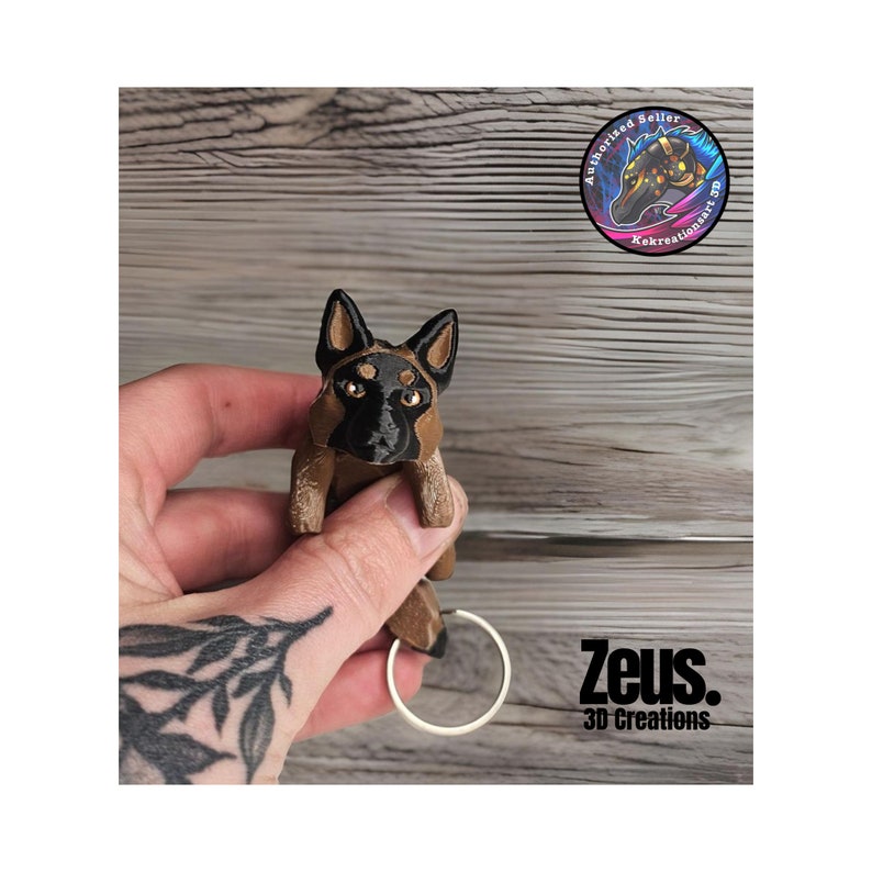 May include: A brown and black German Shepherd dog keychain with a silver ring. The keychain is held in a hand with a tattoo. The text "Zeus. 3D Creations" is visible below the keychain.