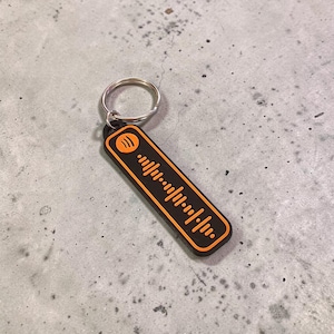 May include: A black and orange rectangular keyring featuring a Spotify code design. The keyring has a silver metal ring attached. The Spotify code is orange and the background is black. The keyring is on a grey surface.