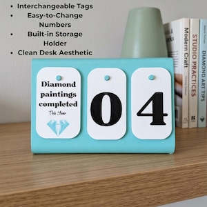Könnte beinhalten: Ein türkisfarbener Schreibtisch-Organizer mit austauschbaren Etiketten mit der Aufschrift "Diamond paintings completed This Year" und der Zahl "04". Der Organizer hat einen eingebauten Aufbewahrungshalter und eine saubere Ästhetik.