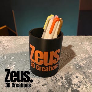 May include: A cylindrical black pen holder featuring the orange text "Zeus 3D Creations" printed on the front. The holder contains several pens with yellow, orange, and brown caps, providing a functional storage solution for writing instruments.